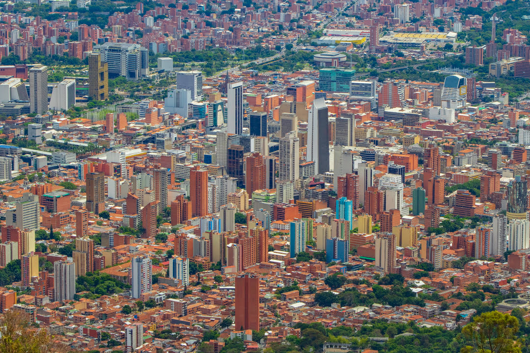 Vista aérea panorámica de la ciudad de Medellín, Colombia, mostrando una alta densidad de edificios. Predominan las construcciones de ladrillo rojo que se mezclan con modernos rascacielos de oficinas y apartamentos de color blanco y gris. Al fondo, se aprecia el relieve montañoso que rodea el valle y algunas estructuras icónicas del centro de la ciudad bajo una luz de día clara.